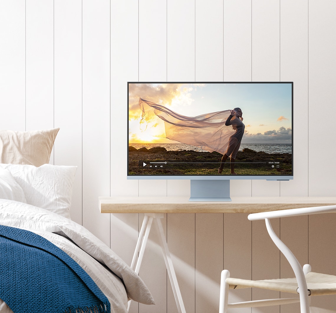 There is a blue monitor on top of a different desk. And a video with a lady standing in the nature is being played on the monitor. A desk chair is shown in front of the desk. To the left of a desk is a bed with pillows and a folded comforter on top.