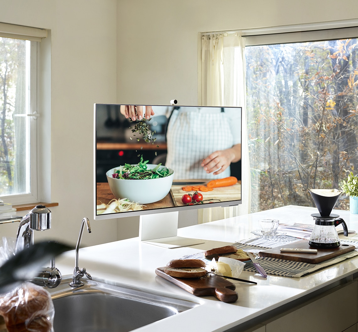 There is a white Smart Monitor image in a kitchen table. A cooking content is being played on the monitor and some kitchen utensils are around it.