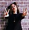 Millie Bobby Brown standing against a brick wall in a black pinstriped blazer and red pants, and forming two peace signs with her hands as she pouts her lips for the camera.