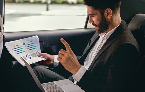 In the backseat of a car, a man in a suit is going through business papers in his hand with a Galaxy Book sitting on his lap.