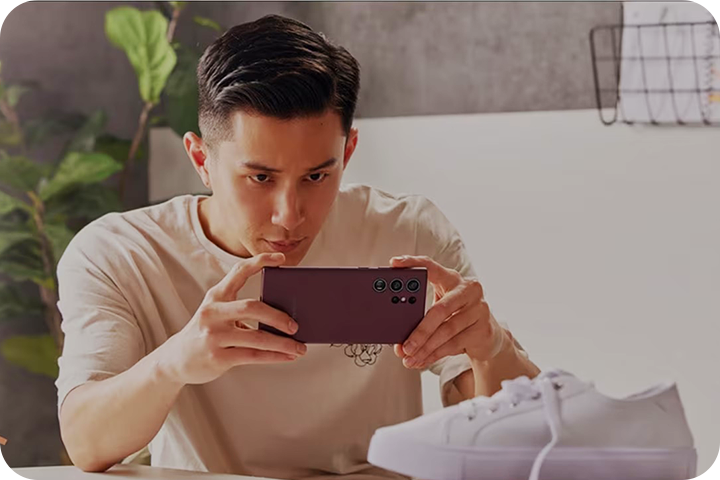 A young man takes a photograph of a shoe with his Samsung device.