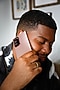 Khalid holds a Mystic Bronze Galaxy Note20 in one hand with its S Pen in the other hand as he laughs. He's sitting on a beige couch and wearing a black and white tie dye t-shirt.