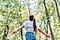 A woman with a long brown ponytail, wearing a white shirt and blue striped shorts stands with her back to the camera. Her head is tipped back and her arms outstretched to the side. She is surrounded by leafy green trees under a blue sky.