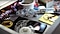 A photograph of a gray desk drawer. The drawer is open, revealing various items within: a roll of Scotch tape, string, measuring tapes, a pad of yellow Post-Its, rings of keys, adapter plugs, cables and a smartphone with a black screen.
