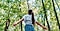 A woman with a long brown ponytail, wearing a white shirt and blue striped shorts stands with her back to the camera. Her head is tipped back and her arms outstretched to the side. She is surrounded by leafy green trees under a blue sky.