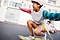 A young woman wearing Galaxy Buds2 rides a skateboard down an empty street. She is wearing round sunglasses, a pink baseball hat, a multicolored striped shirt, blue shorts and checkerboard sneakers. In the background, a young man wearing white sunglasses, a pink shirt and blue shorts sits on a skateboard.