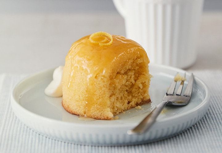A loaf of golden baked sponge cake with lemon zest on top is on a white plate.