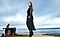 A woman stands by the water in a flowered blouse and a ruffled black skirt. She stands in profile with her hands on her hips in a power pose.