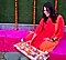 A woman in an orange and red dress sits on a colorful pink striped outdoor cushion. She smiles as she arranges tea lights on a pink tray. Strings of bright orange marigold blossoms are draped behind her against a green turf wall.