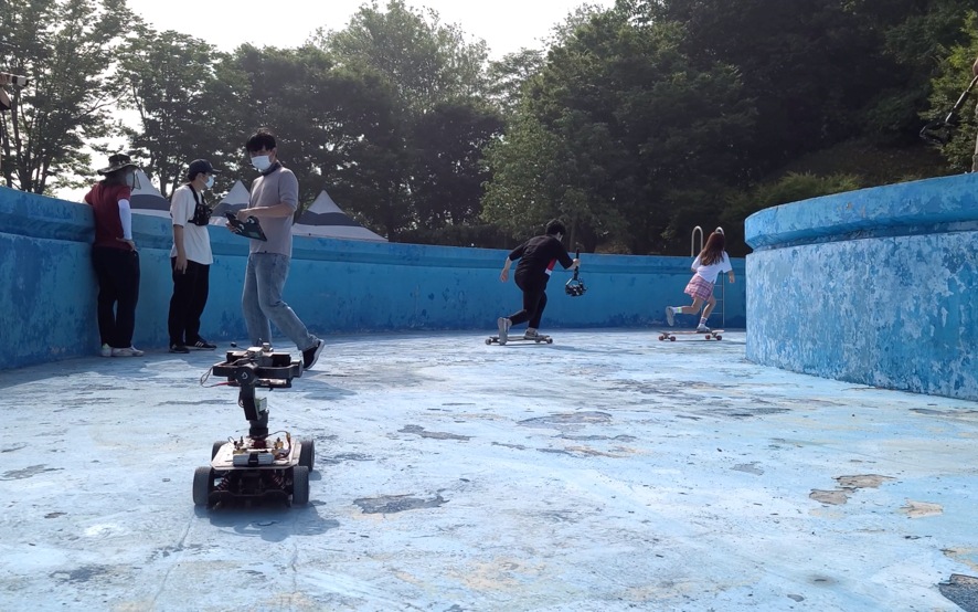 This is a thumbnail for the first how-to video. It shows a mobile remote controlled unit carrying a Galaxy S20 Ultra 5G as it follows an actor on a longboard through an empty pool. Three crew members stand nearby.