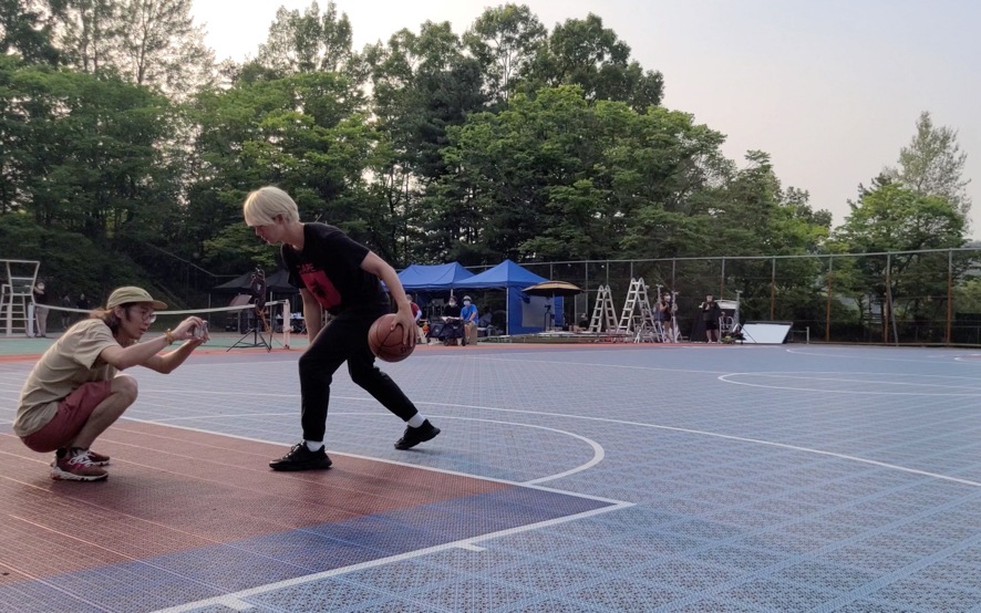 This is a thumbnail for the second how-to video. It shows a crew member filming an actor dribbling a basketball on a basketball court.