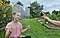 A girl in a pink shirt is sitting on grass, holding a sheet of plexiglass covered with water droplets in front of her face. She's being photographed by someone holding a Galaxy S21 Ultra 5G.