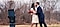 A man in a blue suit (Caleb Natale) kisses a woman in a cream and orange dress outdoors. She is holding a red heart-shaped box. Bare tree trunks and grass are in the background. In the foreground, a Galaxy S22 Ultra is mounted vertically on a tripod. The screen shows an image of the couple captured through the phone's camera lens.