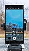 A Galaxy S22 Ultra is mounted vertically on a tripod outdoors. On the screen is an image of video creator Caleb Natale wearing a blue, gray and black hoodie and black pants. The camera controls appear on the screen over the image and there is a filter on the image saturating the colors in contrast to the background.