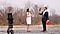 A man in a blue shirt suit (Caleb Natale) stands outside presenting a bouquet of red roses and a red heart-shaped box to a woman in a cream and orange dress. There are bare trees, grass and a gravel road in the background. In the foreground, In the foreground, a Galaxy S22 Ultra is mounted vertically on a tripod. The screen shows an image of the couple captured through the phone's camera lens.
