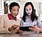 A young girl and her grandmother sit next to each other at a kitchen counter. The young girl holds a black Galaxy tablet and looks at the screen. The grandmother looks on and points to something on the screen.