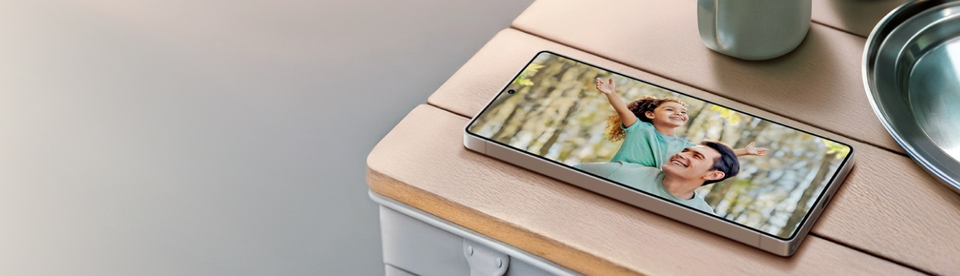 A Galaxy smartphone lies on a wooden table displaying a photo of a father and child. Next to it are a mug, a moka pot, and a metal plate. The background includes soft morning light and the headline “Discover Father’s Day gifts” with a “Shop the gift guide” button.