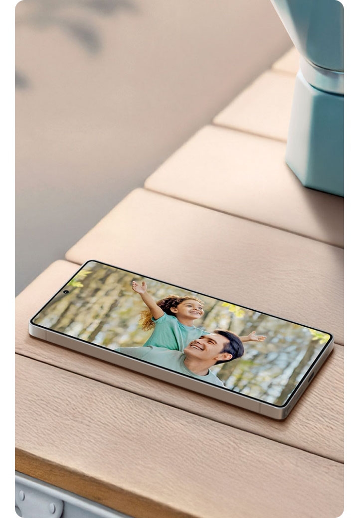A Galaxy smartphone lies on a wooden table displaying a photo of a father and child. Next to it are a mug, a moka pot, and a metal plate. The background includes soft morning light and the headline “Discover Father’s Day gifts” with a “Shop the gift guide” button.