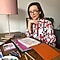 A short-haired woman with glasses sitting at her desk smiling wearing a white blouse with a red and blue pattern. On her desk are art materials and there is a pink lamp in the background.