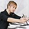 A light-haired woman in a black top is sitting and painting a large piece of art. She is holding a brush on her right hand and touching the canvas to paint with a black ink.