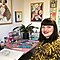 A short-haired woman with dark hair is sitting at her desk smiling wearing a leopard print top. On her desk are art materials and there are colourful art prints in the background.