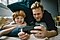 Father and son lay in bed on their bellies as the father holds a dark grey smartphone in landscape mode. Both the father and son smile as they gaze into the phone's display as if they're watching a show at home.