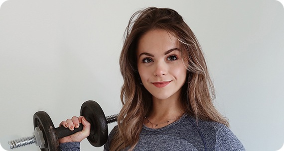 A woman in a sports attire is shown. She is holding a weight in her right hand, inside a room with a white door. She is looking content, with a subtle smile on her face.