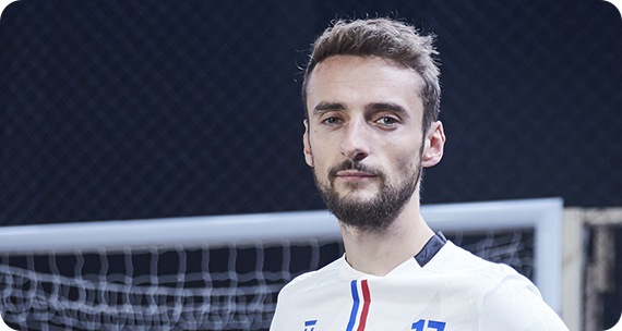 A man in a soccer jersey is shown. He is standing with his hands on his waist on a soccer field.