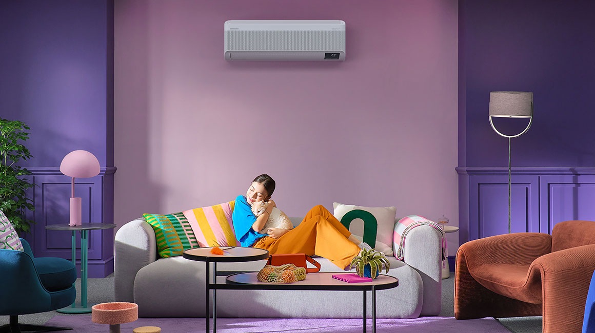 Một cô gái trẻ đang ôm một con mèo khi cô ấy đang thư giãn trên ghế couch. Đằng sau cô ấy là điều hòa treo tường WindFree