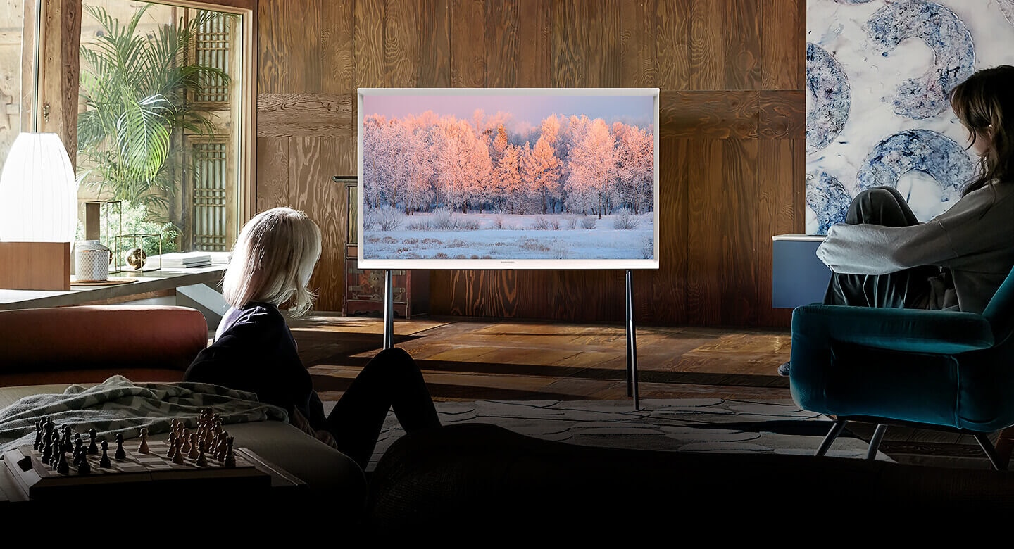 Hai người phụ nữ đang nhìn TV The Serif màu Cloud White được dựng trên chân đế trong một phòng khách thanh lịch. Màn hình The Serif đang hiển thị một cảnh mùa đông có tuyết trắng và rừng cây. Và đang phóng to vào trong để tập trung sự chú ý đến The Serif.