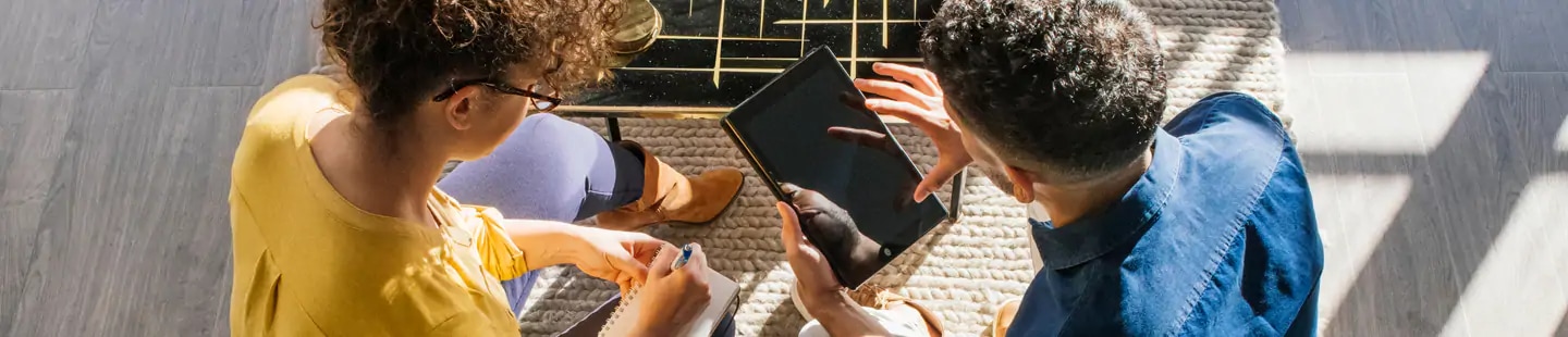 Hình quay từ trên cao một thanh niên đang cầm máy tính bảng và một cô gái đang cầm notepad và bút. Người thanh niên dường như đang giải thích điều gì đó với cô gái.
