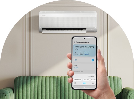 The WindFree AC is mounted on a white wall behind a green sofa. A hand holds a smartphone, displaying the “Room Air Conditioner” screen.