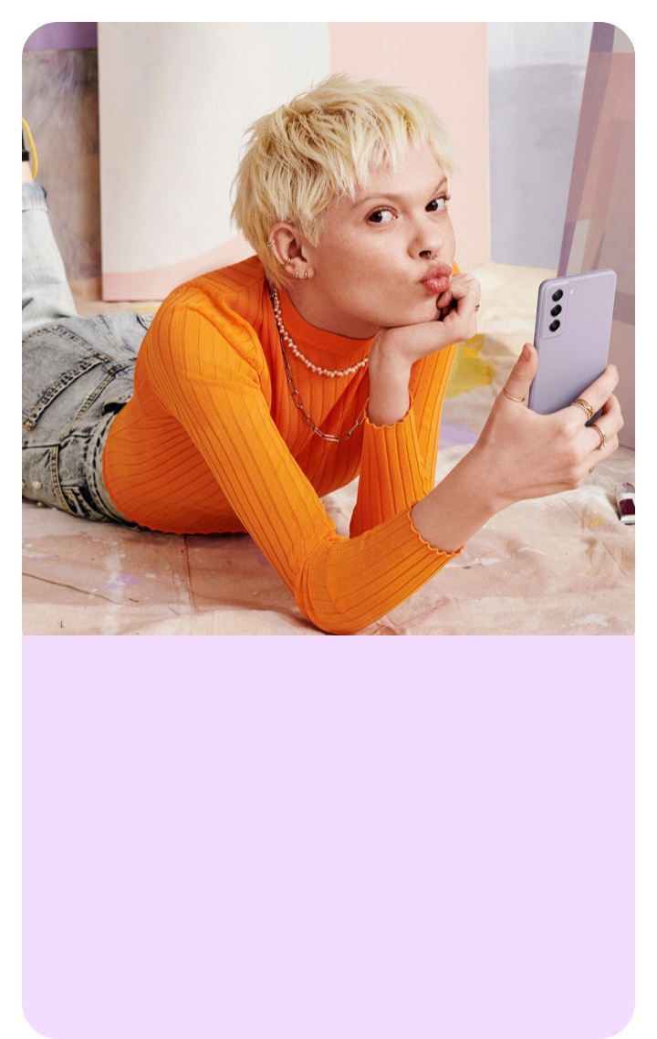 Une femme allongée par terre avec un Galaxy S22. Elle mime un baiser et est entourée de ses tableaux et peintures.