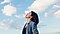 A woman in a blue-gray blazer stands with her head tilted toward a blue sky with white puffy clouds. Her eyes are closed and she looks to be inhaling deeply.