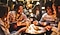 A group of four adults sitting outdoors around a tabletop fire pit roasting marshmallows.