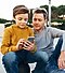 An outdoor scene of a young boy sitting on a basketball and looking at his phone with his father sat beside him