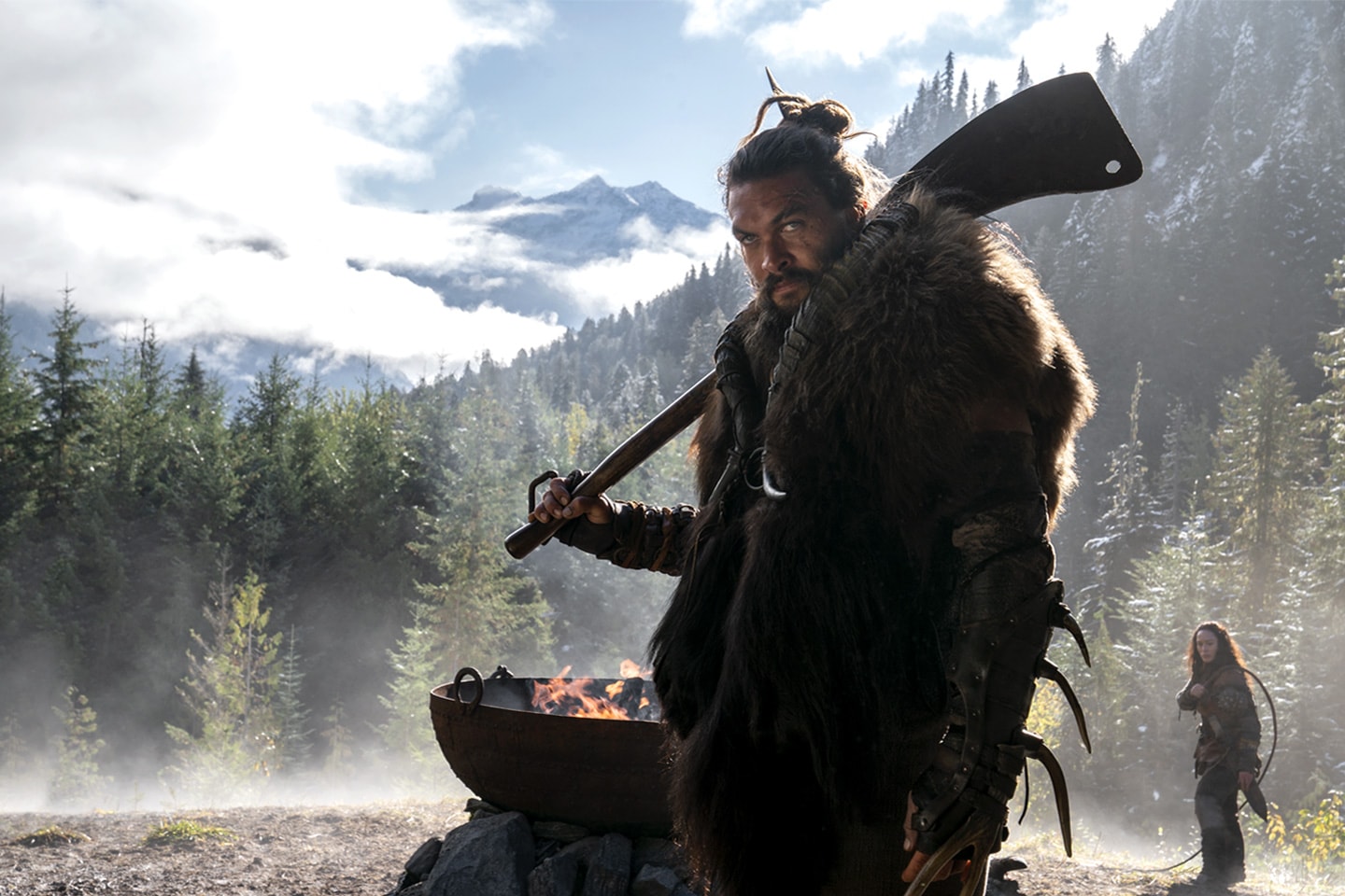 A man in the mountains stands with a weapon in front of a fire pit with a woman behind him.