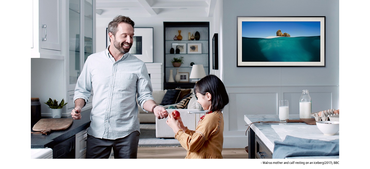A man and his child are together in the kitchen. The Frame is on the wall displaying 'Walrus mother and calf resting on an iceberg (2017)' by BBC, a picture of a walrus and her calf on top of an iceberg in the deep turquoise sea.