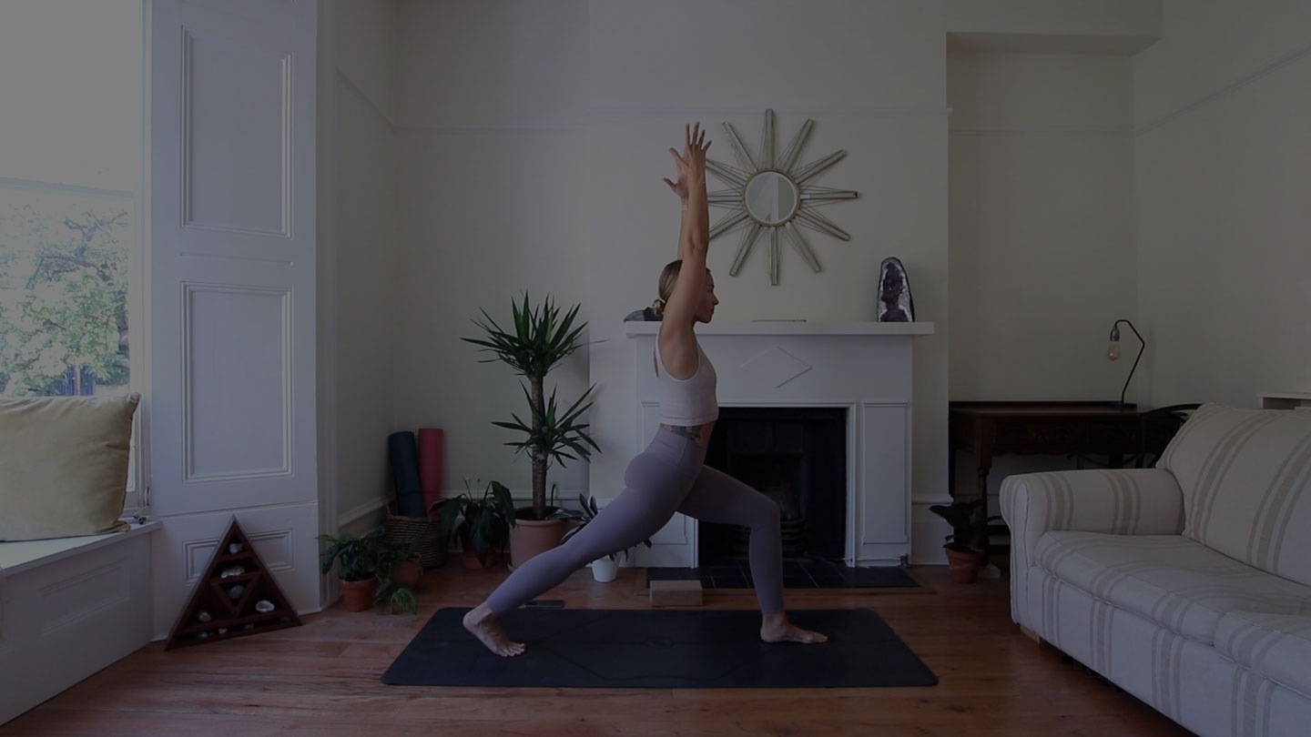 Cat Meffan in a Warrior I pose on a black yoga mat in a bright living room