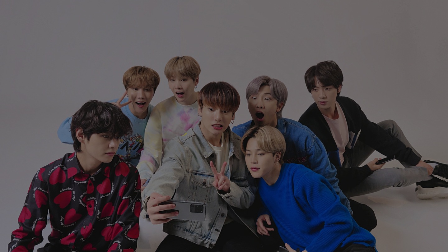BTS taking Group Selfie. All members are sitting on the ground and taking a BTS selfie on a Galaxy S20+ held by Jung Kook.