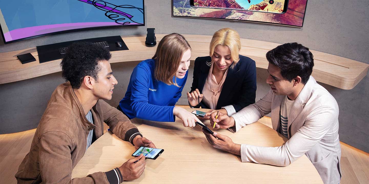 A woman and two men are sitting around a desk with the Galaxy Consultant at their Galaxy Workshop event with their smart phones in their hands. There are monitors and keyboard in the background.