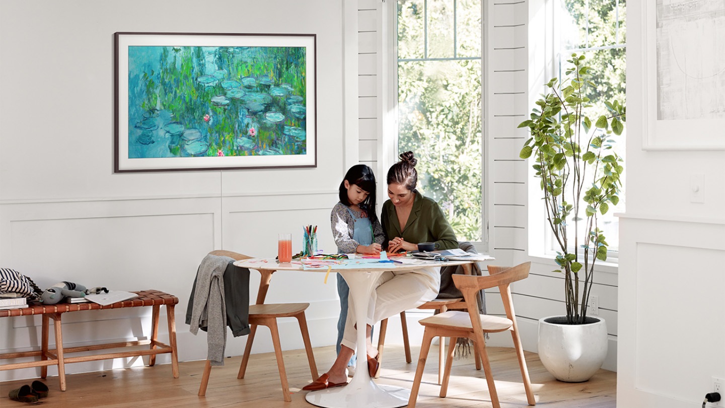 gallery image 5 The Frame with a black bezel is mounted stylishly on a white wood wall in a kitchen. Onscreen is an image of water lilies on a wall and in the foreground a mother and daughter are drawing something at a table.