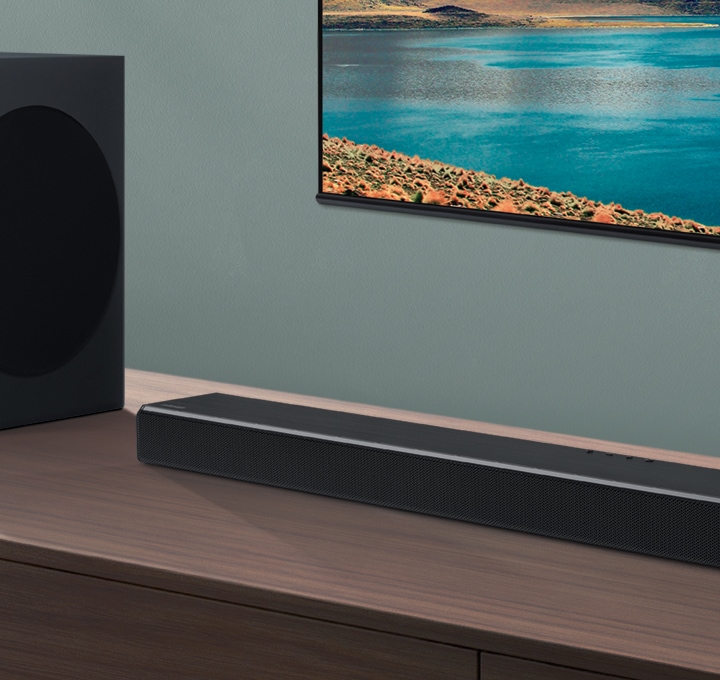 Soundbar and subwoofer are seen on wood cabinets at an angle, with a TV mounted on a wall in the background. Soundbar and subwoofer are seen on wood cabinets at an angle, with a TV mounted on a wall in the background.