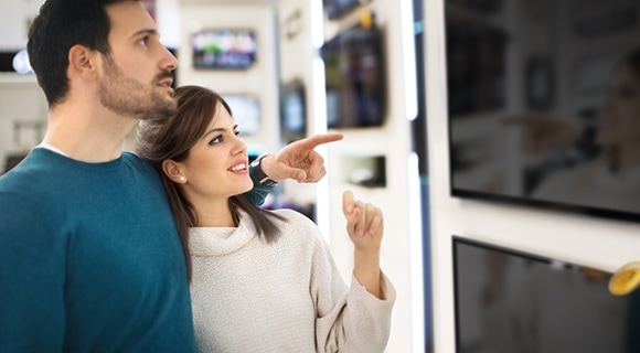 A couple stands and points at big screen TVs while shopping.