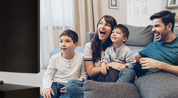 A laughing and smiling family of four are sitting closely together on a sofa enjoying a big screen TV.