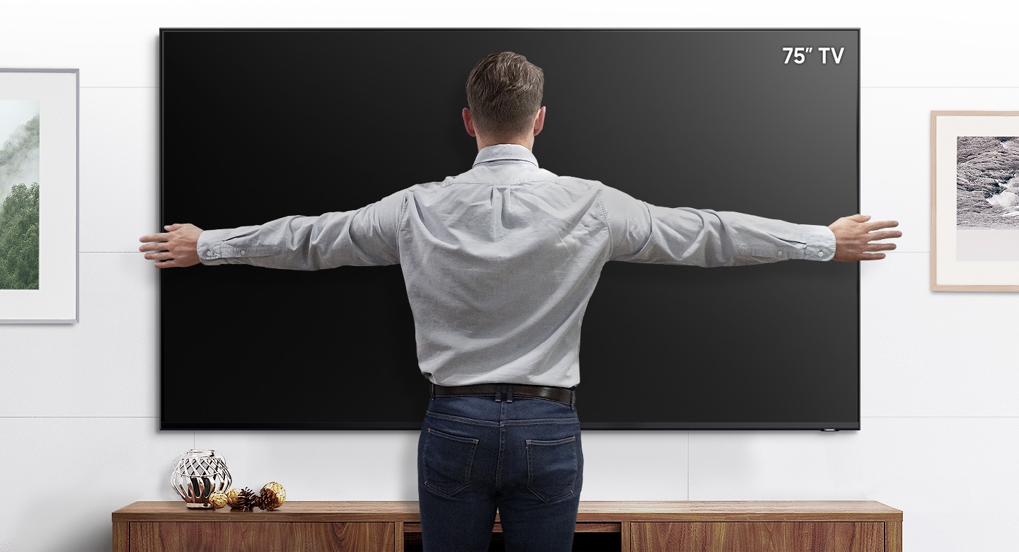 A man stands in front of a wall mounted 75” TV with his arms open to show that the TV fits within his arm span.