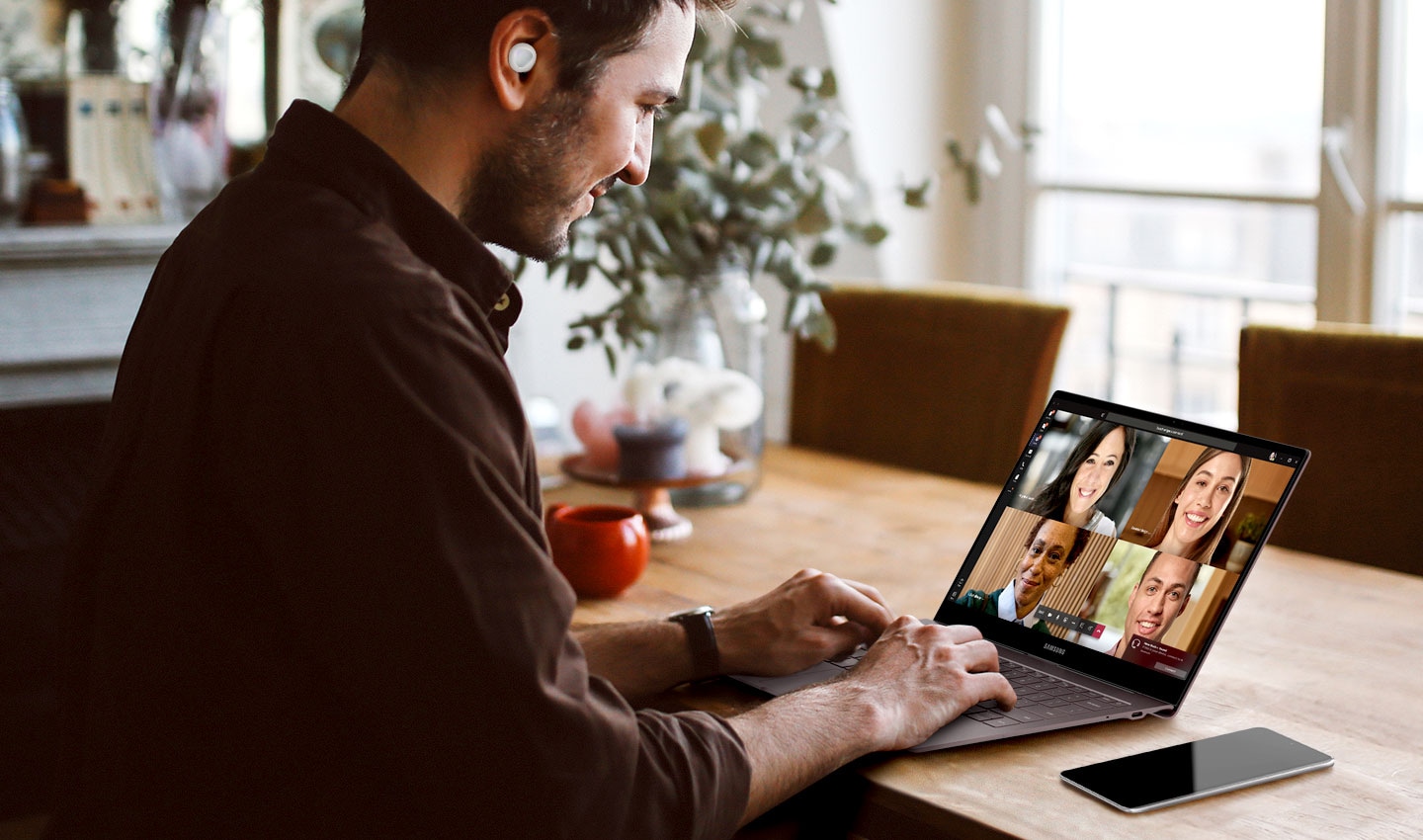 Man sits at home wearing white Galaxy Buds plus earbuds as he participates in a team conference call on his Windows PC.