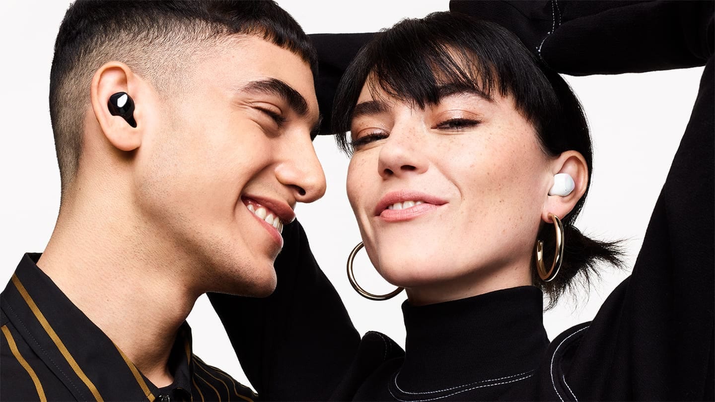 A man wearing black Galaxy Buds plus smiles as he looks a woman next to him wearing white Galaxy Buds plus. The woman smiles with her hands in the air behind her head.