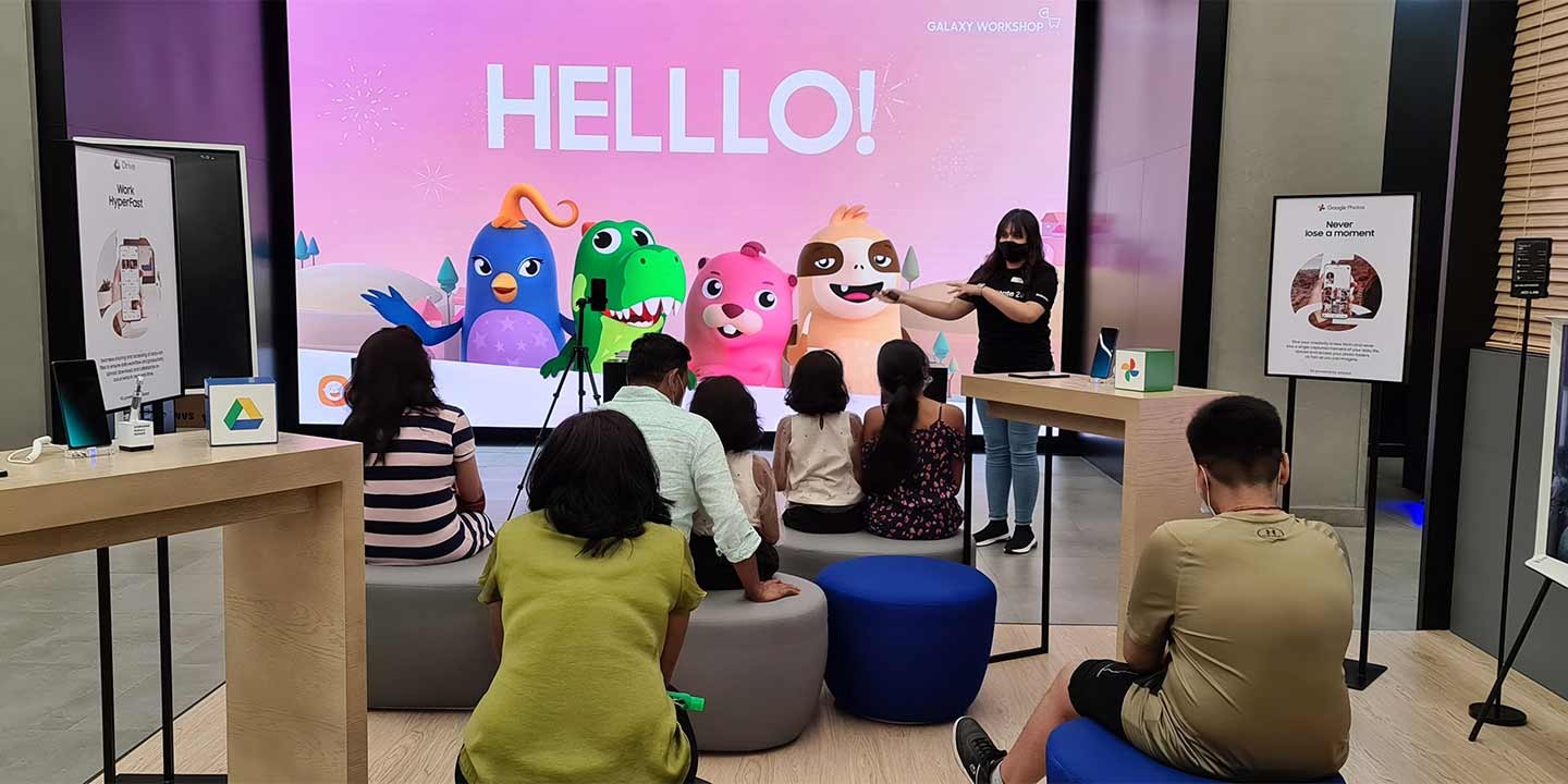 A woman and two men are sitting around a desk with the Galaxy Consultant at their Galaxy Workshop event with their smart phones in their hands. There are monitors and keyboard in the background.