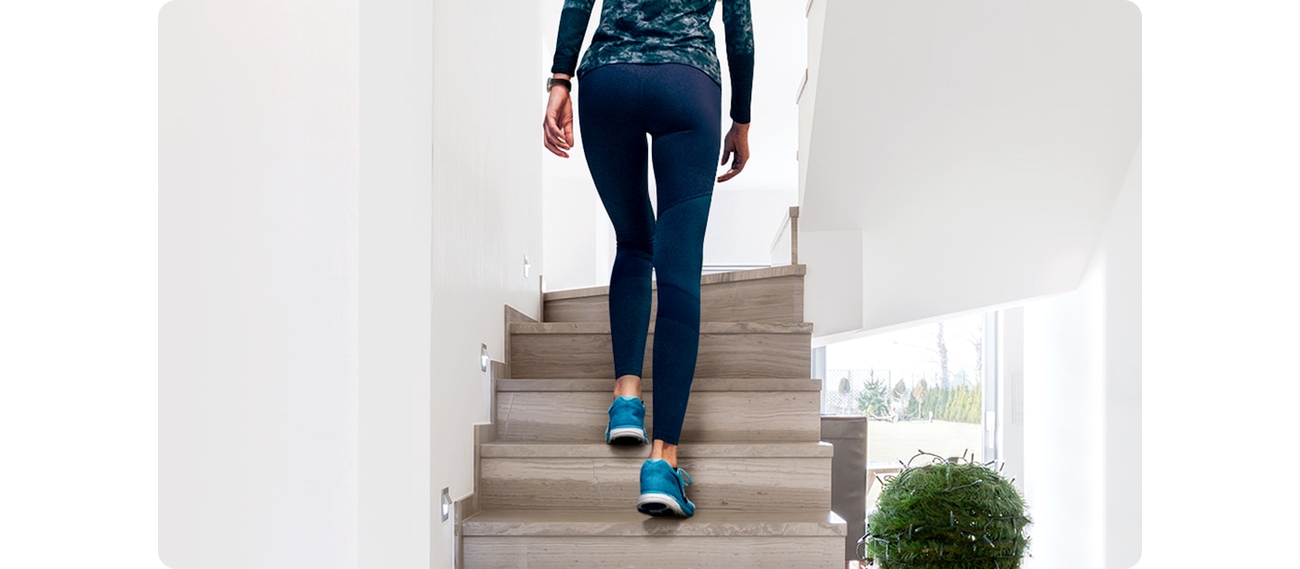 A woman climbs an indoor staircase. She wears athletic clothing and sneakers, with a Galaxy Watch on her wrist.
